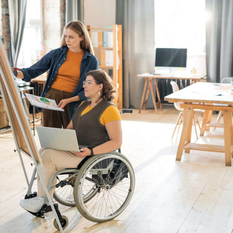 Young businesswoman in casualwear helping her disable colleague with presentation while putting papers with points on whiteboard in office